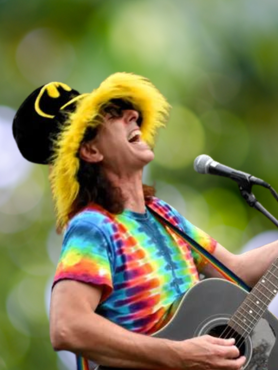 A tall man with shoulder-length dark hair wears tie-dye and an ostentatious hat while playing guitar.