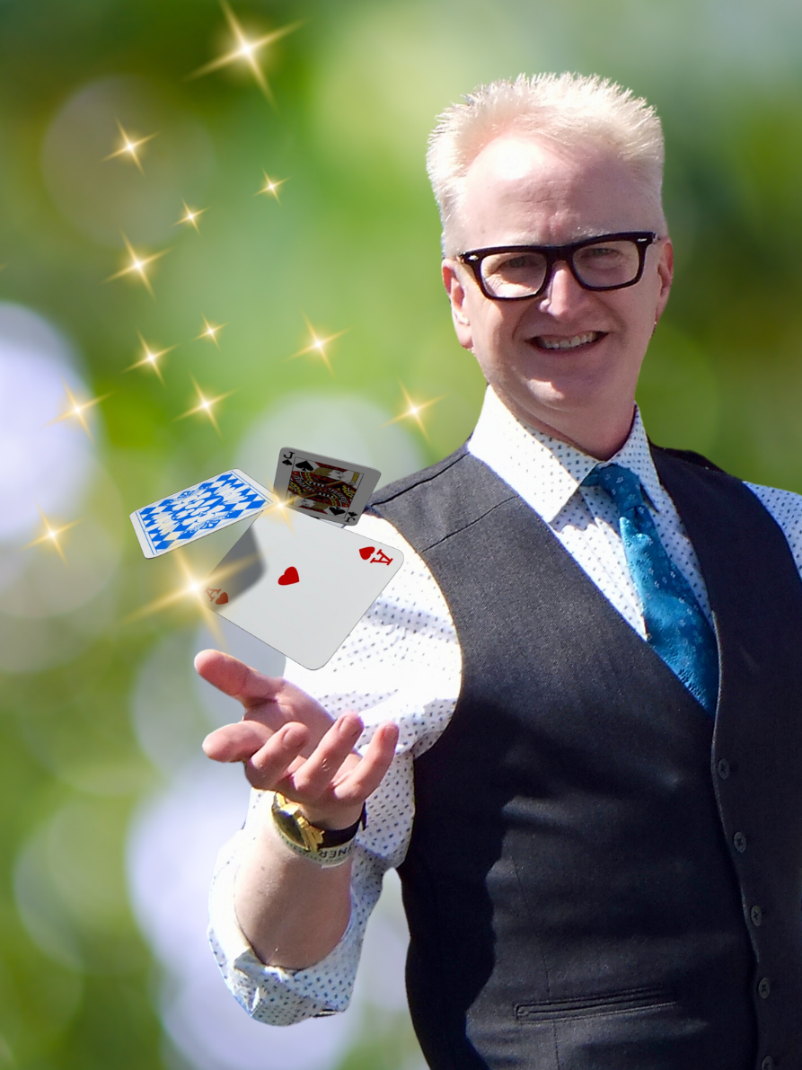 A man wearing a suit vest, dark glasses, and sporting a shock of short, white-blonde hair does magic tricks with playing cards.