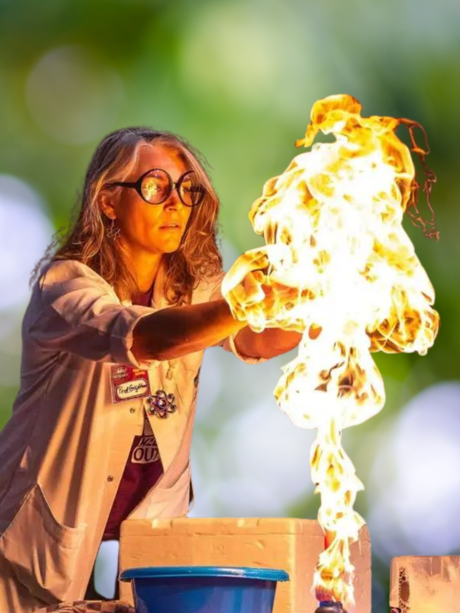 A woman with long, blonde hair wearing a lab coat and protective glasses does an experiment producing a controlled, fiery explosion!