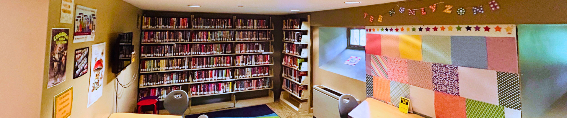 A view of the Teen Den in September of 2025 featuring a blue and green rug, shelves of books, and a colorful patchwork wall with a banner reading "TEEN ONLY ZONE" hanging above it.