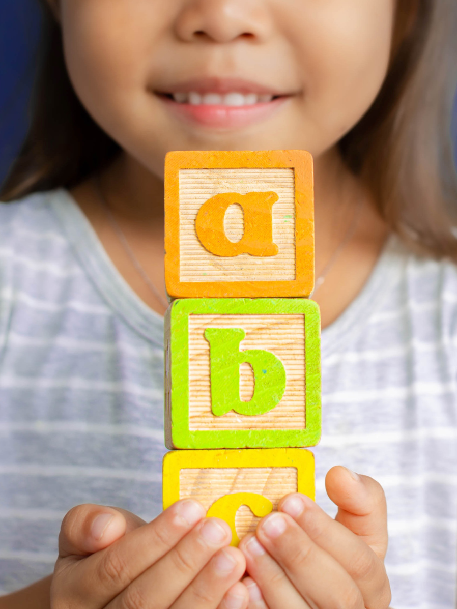 A smiling child holds a stack of blocks showing the first three letters of the alphabet.