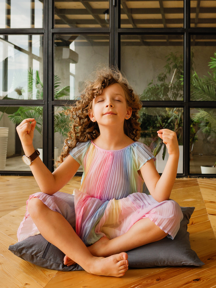 A peaceful-looking, curly-haired child in a pastel rainbow dress practices a seated yoga pose.