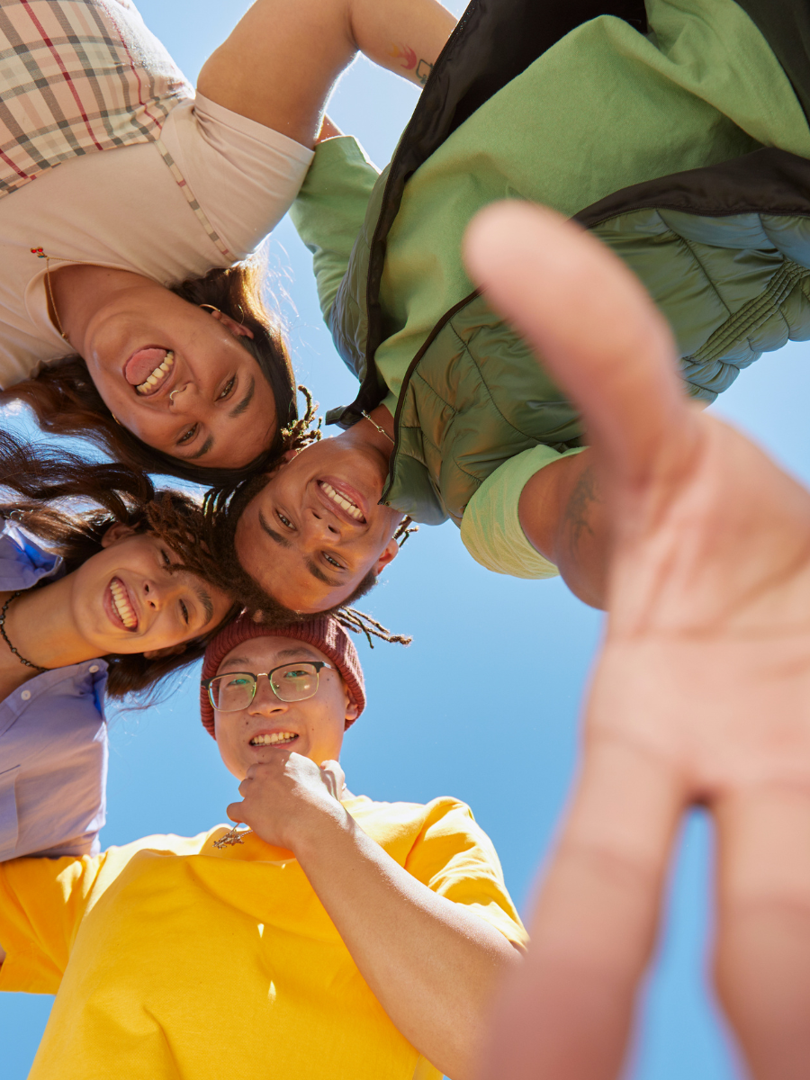 A group of smiling teens in a huddle, looking down at the camera, with a blue background. One teen has a hand outstretched toward the camera. 