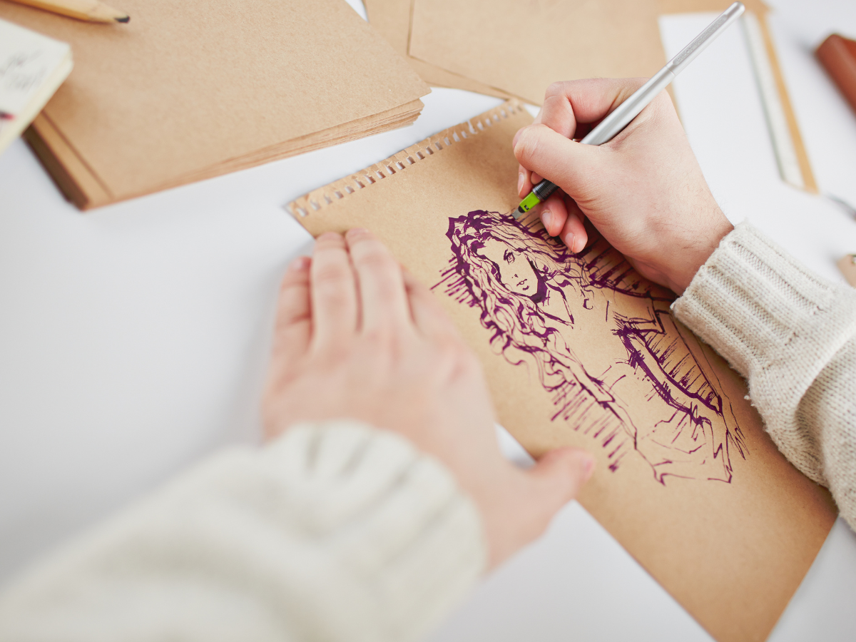 Hands holding a pen and drawing a girl on brown paper with purple ink.