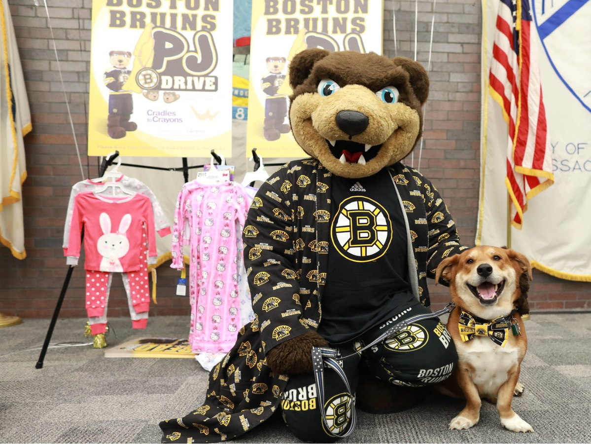 Blades the Bruins bear wearing pajamas, posing with a happy dog in front of donated pajamas.