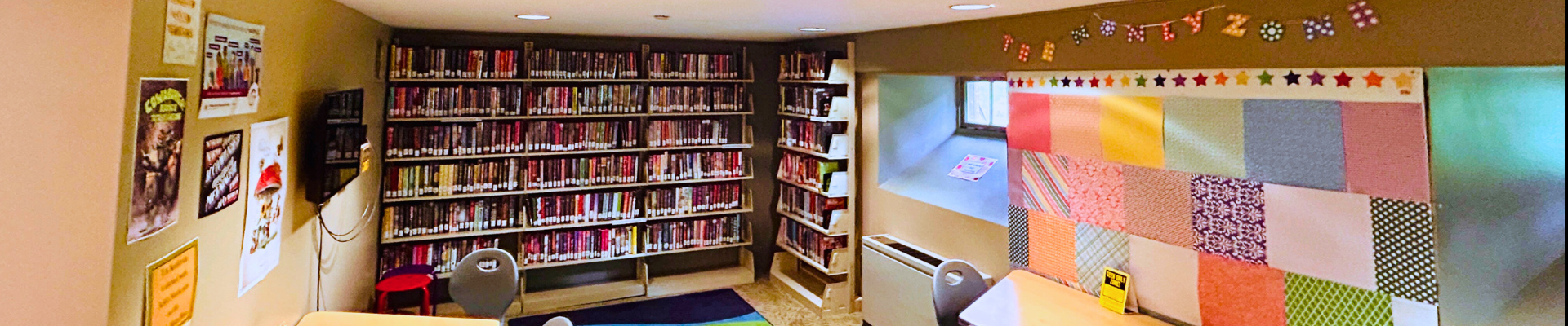 A view of the Teen Den in September of 2025 featuring a blue and green rug, shelves of books, and a colorful patchwork wall with a banner reading "TEEN ONLY ZONE" hanging above it.